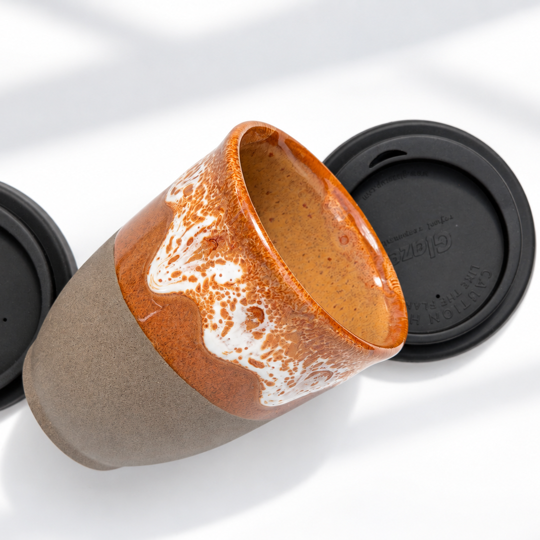 A ceramic reusable coffee mug with a brown and orange glazed upper half and a matte taupe-brown lower half, shown with a black lid on a white background.