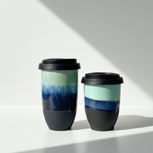 Two reusable travel mugs with black lids and black lower sections, featuring a blue and mint green gradient design on the upper body, photographed on a white surface with a diagonal shadow.