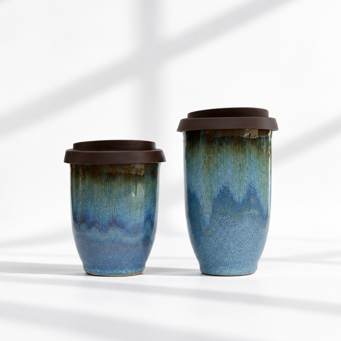 Two ceramic reusable travel mugs with dark brown lids, featuring a blue and green glaze with a dripping effect, set against a white background with soft diagonal shadows.