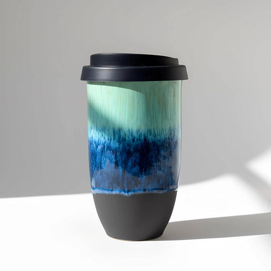 A ceramic travel tumbler with a black lid, featuring a gradient design that transitions from mint green and teal at the top to deep blue in the middle, and finishes with a matte black base.