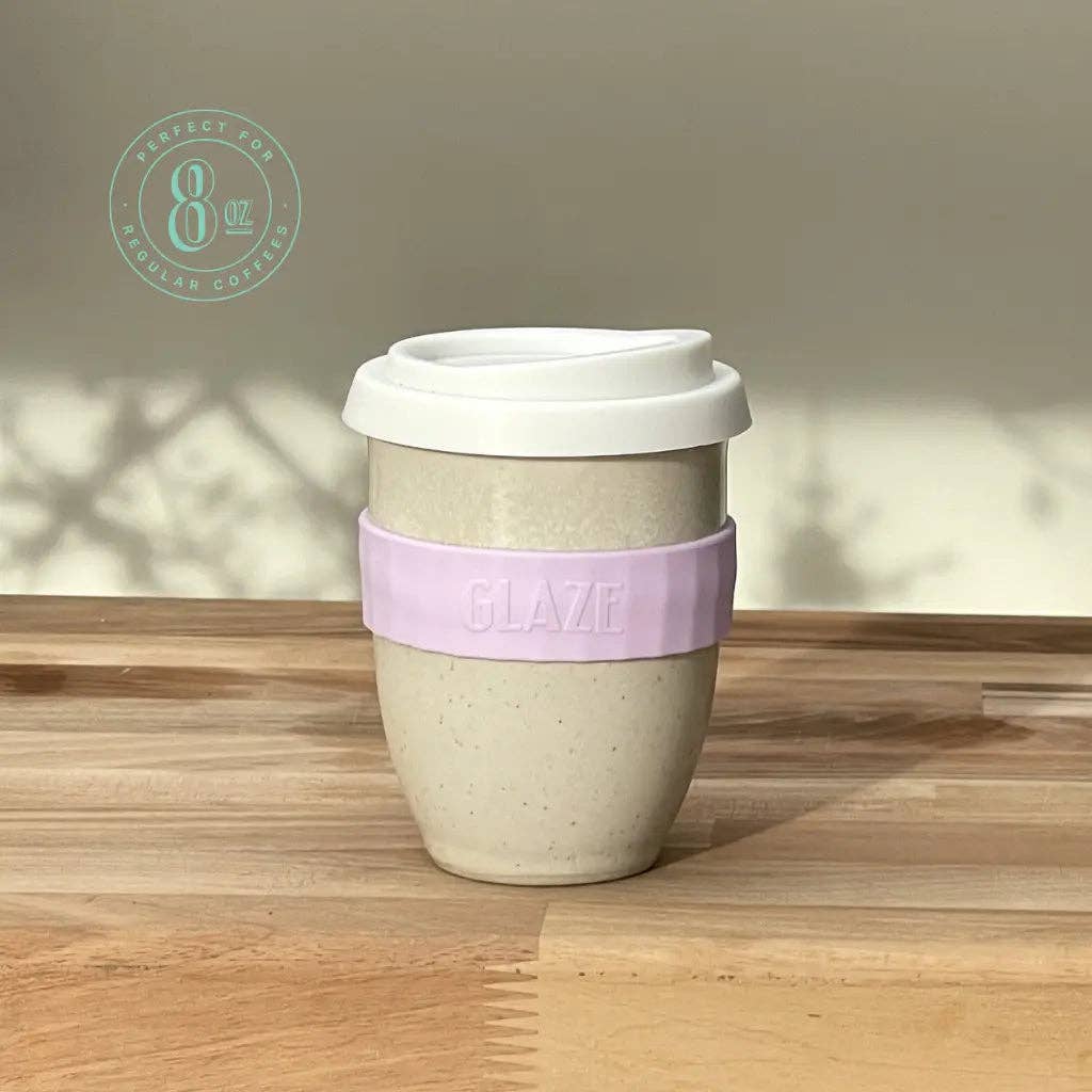 An 8 oz reusable coffee cup with a white lid, speckled beige body, and a lavender silicone sleeve embossed with the brand name 'GLAZE'. The cup is placed on a wooden tabletop with soft shadows cast on the wall behind it.