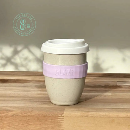 An 8 oz reusable coffee cup with a white lid, speckled beige body, and a lavender silicone sleeve embossed with the brand name 'GLAZE'. The cup is placed on a wooden tabletop with soft shadows cast on the wall behind it.