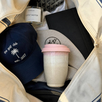 A curated collection of travel and lifestyle accessories packed inside a cream-colored zippered bag, including a navy blue baseball cap with white embroidery, a pink-tipped reusable coffee cup, a black notebook, a glass bottle of Sypsy Water, and a gray patterned necktie.