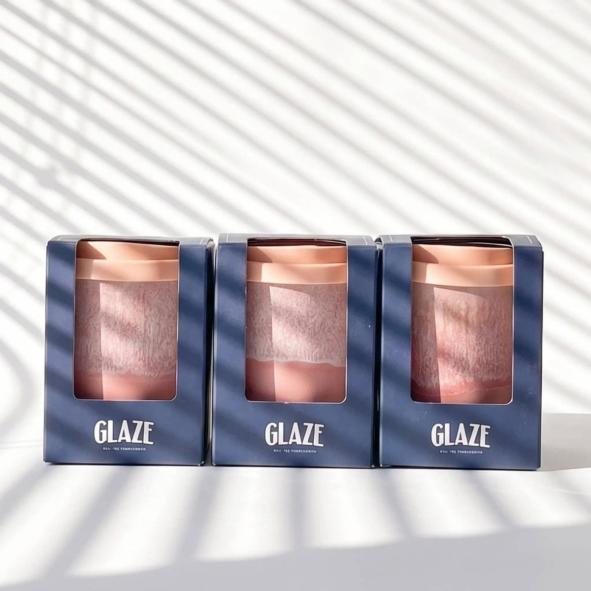 Three GLAZE reusable travel mugs in dark blue packaging, arranged side by side on a white surface with diagonal windowpane shadows.