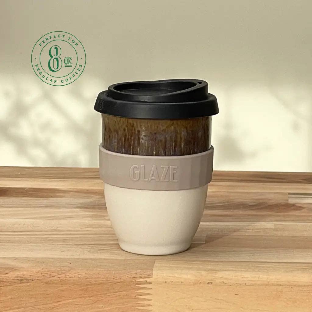 An 8 oz reusable coffee cup with a black lid, a dark brown wood-grain textured middle section, and a beige silicone sleeve embossed with the brand name 'GLAZE'. The cup sits on a light wooden tabletop against a neutral beige wall with soft leaf shadows.