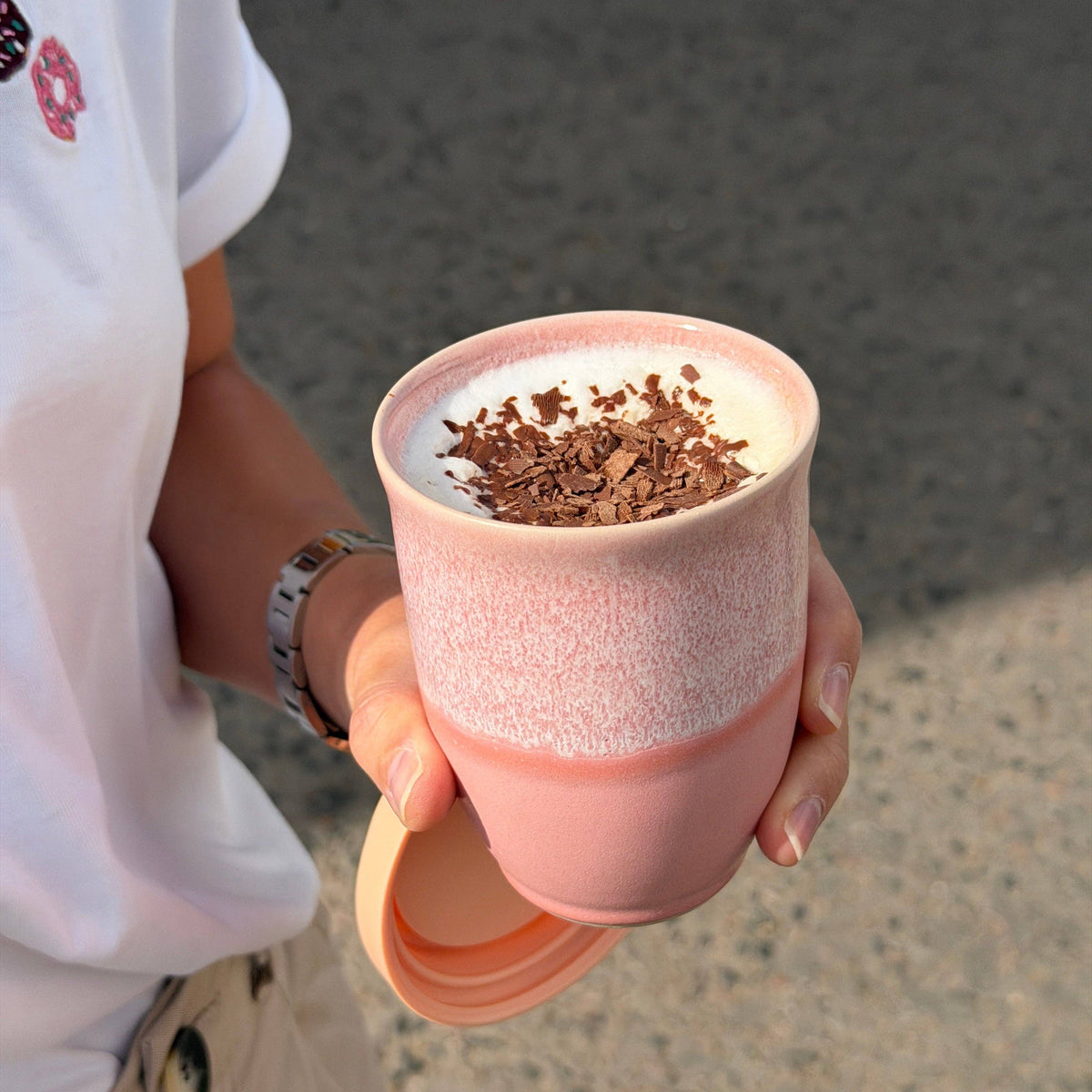 A person holds a pink ceramic travel mug with a matching lid outdoors. The mug has a speckled pink upper section, a smooth pink lower section, and a white layer of foam on top with chocolate shavings sprinkled over it.