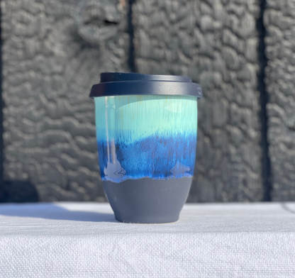 A ceramic travel tumbler with a blue and mint green gradient body, a dark gray base, and a black lid, sitting on a white textured fabric surface with a dark wooden background.