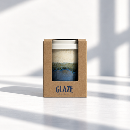 A reusable ceramic mug with a white lid, featuring a blue and green glaze pattern, presented in its brown kraft paper packaging box with the brand name 'GLAZE' printed on the front, set against a bright white background with soft window shadows.