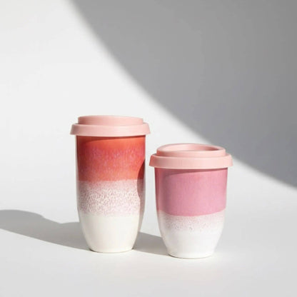 Two reusable travel mugs with pink lids, featuring a gradient design that fades from coral and pink at the top to white at the bottom, set against a light gray background with soft shadows.