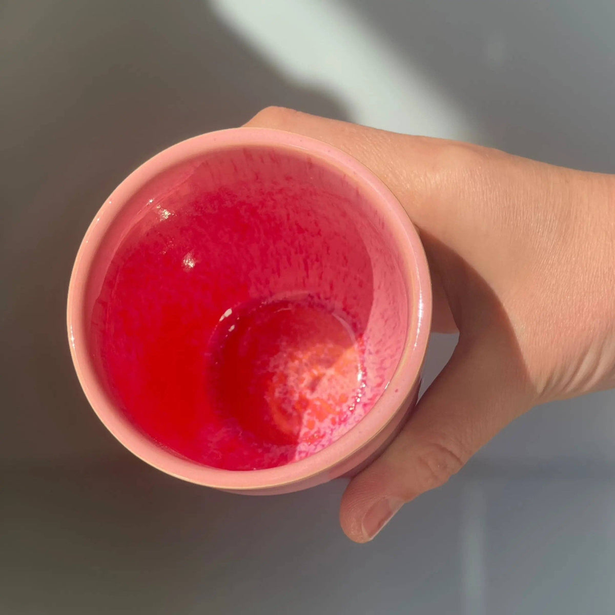 A hand holding a small pink ceramic cup with a vibrant red glazed interior, shown from a top-down angle against a neutral gray background.