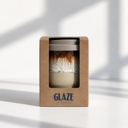 A ceramic reusable travel mug with a brown and white marbled glaze and a gray lid, presented inside a brown kraft paper window box with the brand name 'GLAZE' printed in blue at the bottom.