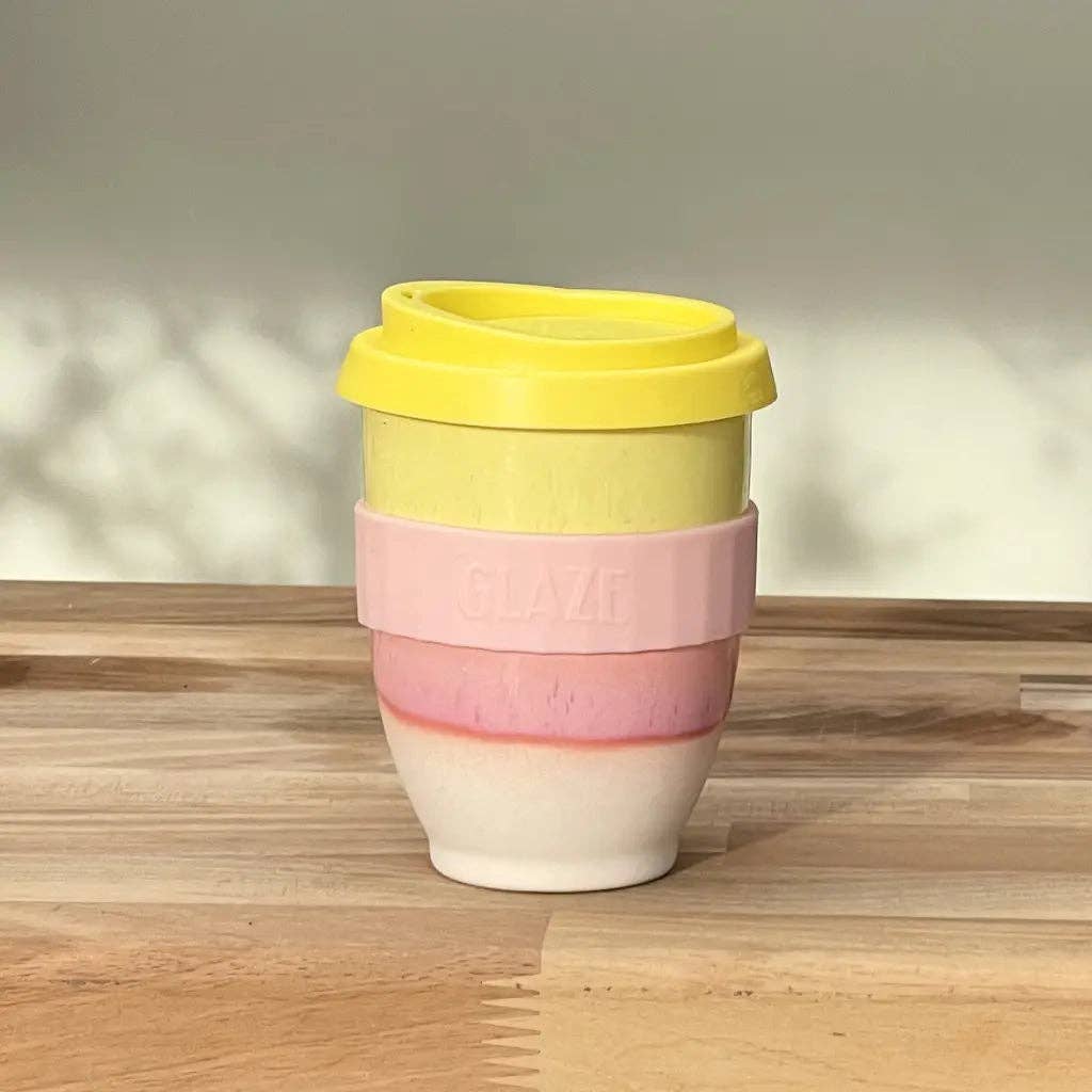 A reusable travel tumbler with a yellow lid, a light pink silicone band embossed with 'SLAVI', and a white ceramic lower section, shown on a wooden tabletop against a neutral wall.