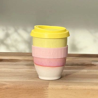 A reusable travel tumbler with a yellow lid, a light pink silicone band embossed with 'SLAVI', and a white ceramic lower section, shown on a wooden tabletop against a neutral wall.