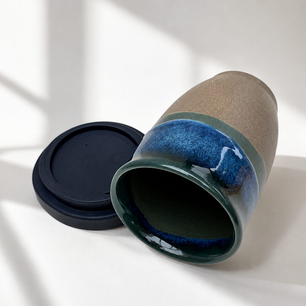 A ceramic travel mug with a dark blue and green glazed interior and a matte taupe-brown exterior, shown with its black lid on a white surface with soft window shadows.