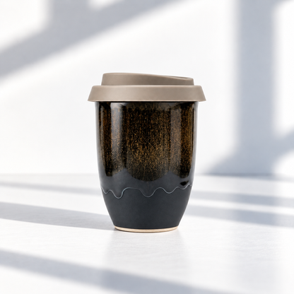 A reusable ceramic travel mug with a dark brown and black speckled glaze, a wavy lower band, and a matte taupe lid, set on a white surface with soft window shadows.