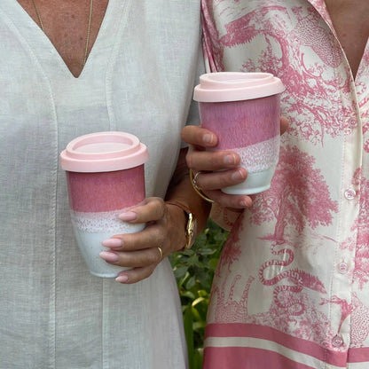 Two reusable ceramic travel mugs with pink lids are held by two women outdoors. The mug on the left is solid dusty pink, and the mug on the right has a pink-to-white gradient with a speckled middle band.