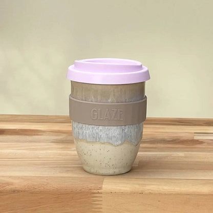 A reusable ceramic coffee cup with a lavender lid, a gray silicone sleeve embossed with the brand name 'GLAZE', and a two-toned beige and blue glazed ceramic body, set on a wooden tabletop against a soft beige background.
