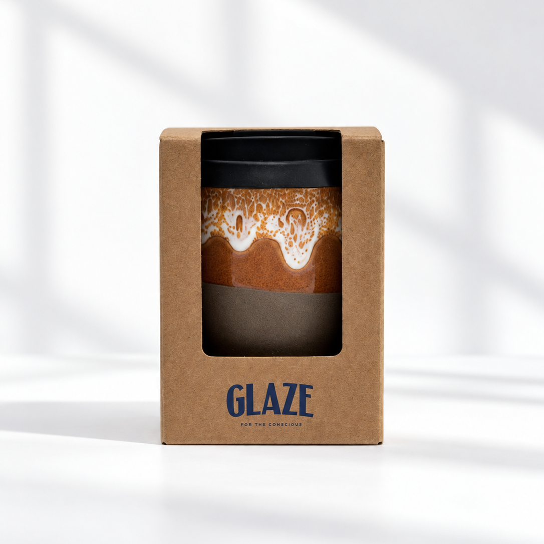 A GLAZE branded kraft paper box containing a reusable coffee cup with a black lid and a brown sleeve featuring a latte art design, photographed on a clean white surface with soft window shadows.