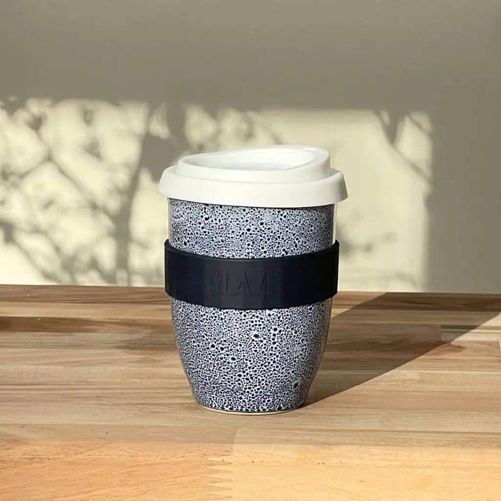 A reusable ceramic travel mug with a speckled gray and white pattern, a black silicone sleeve, and a white lid, sitting on a wooden table with dappled sunlight and shadows.