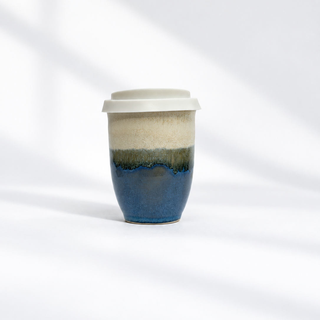 A ceramic travel tumbler with a white lid, featuring a hand-painted glaze with horizontal bands of cream, olive green, and deep blue, set against a white background with soft diagonal shadows.