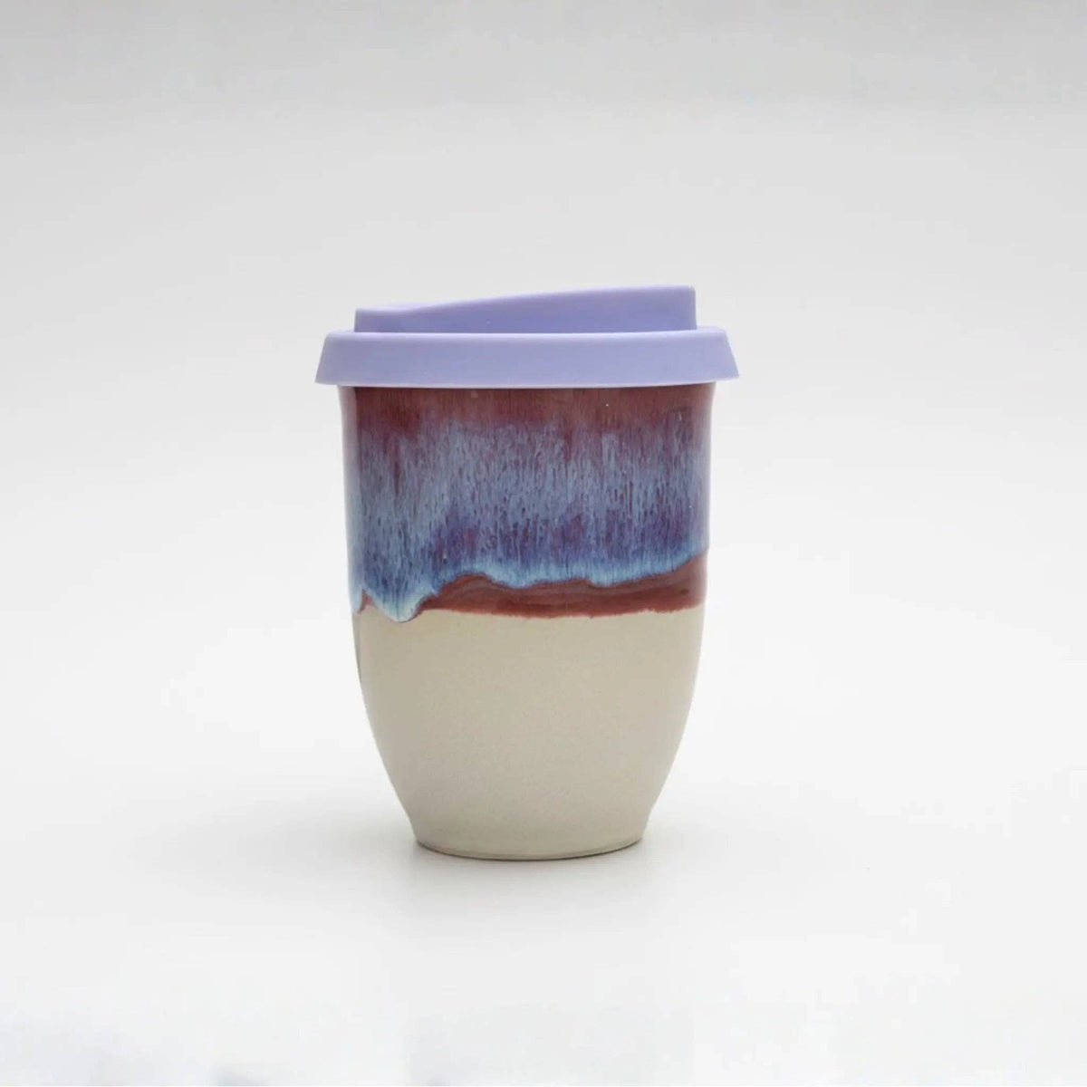 A ceramic travel tumbler with a light purple lid, featuring a two-tone glazed body with a cream-colored lower half and a blue-to-purple marbled upper half.