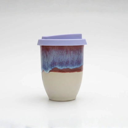 A ceramic travel tumbler with a light purple lid, featuring a two-tone glazed body with a cream-colored lower half and a blue-to-purple marbled upper half.
