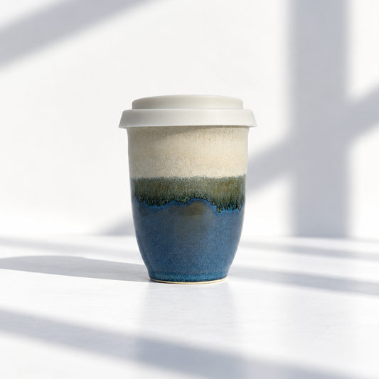A ceramic travel tumbler with a white lid, featuring a three-layer glaze design with cream, olive green, and deep blue sections, set against a bright white background with soft window shadows.
