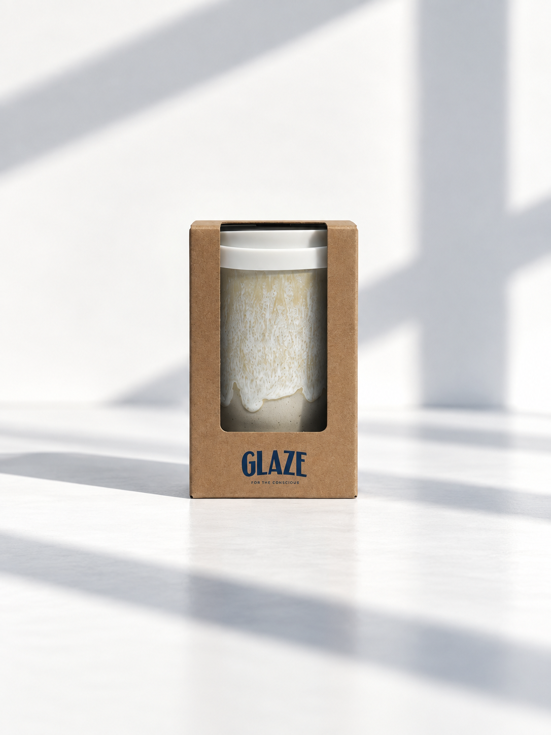 A ceramic mug with a white and beige textured glaze, presented in its brown kraft paper packaging box with the brand name 'GLAZE' printed in blue. The scene is set on a white surface with soft, diagonal window light shadows.