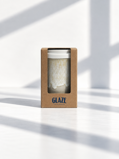 A ceramic mug with a white and beige textured glaze, presented in its brown kraft paper packaging box with the brand name 'GLAZE' printed in blue. The scene is set on a white surface with soft, diagonal window light shadows.