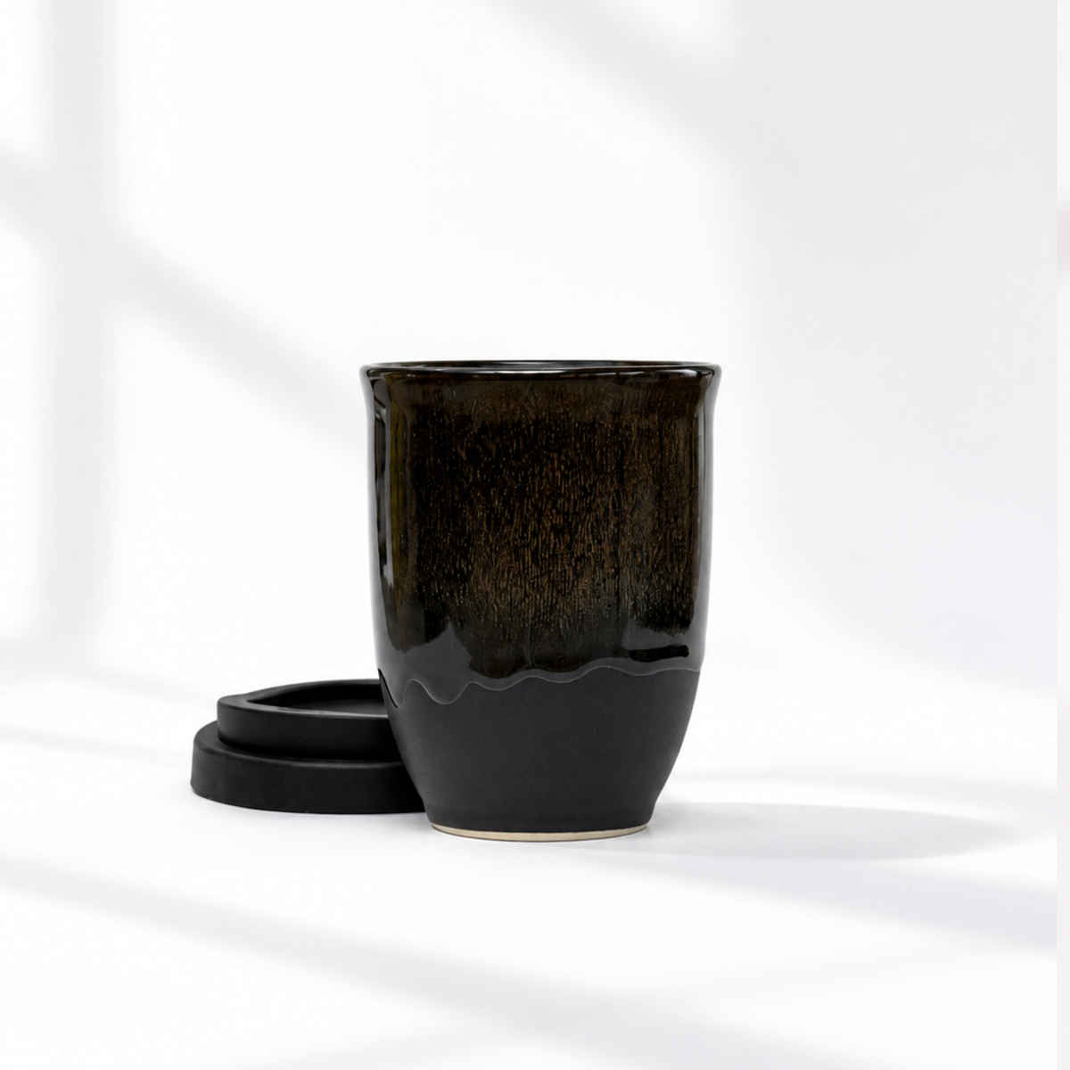 A glossy ceramic tea cup in a dark brown to black gradient glaze, shown with its matching black lid on a clean white surface with soft window shadows.