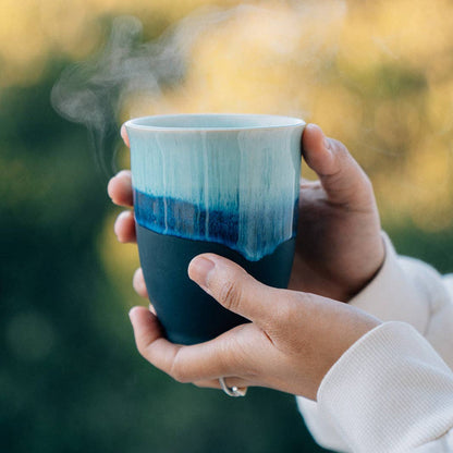 A person holds a steaming ceramic mug with a white upper section, blue gradient middle, and dark navy lower half against a blurred outdoor background.