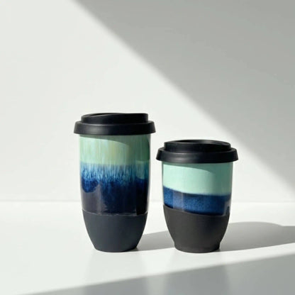 Two reusable travel tumbler cups with black lids and black silicone bases, featuring a glossy ceramic body with a blue and mint green gradient design, set against a white background with diagonal shadows.