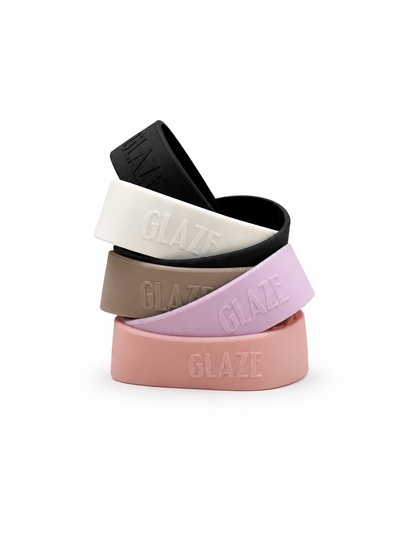 Four silicone bands in black, white, taupe, and pastel pink, stacked on a white background. Each band features the brand name 'GLAZE' embossed on the front.