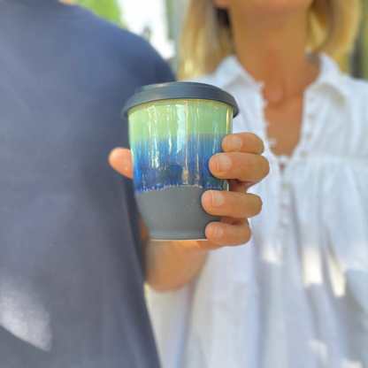 A reusable insulated tumbler cup with a dark gray lid and base, featuring a green-to-blue gradient body, held by a person wearing a white blouse.