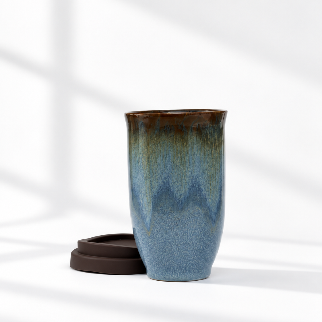 A ceramic travel mug with a blue and green glaze, shown next to its dark brown silicone lid on a white surface with soft window shadows.