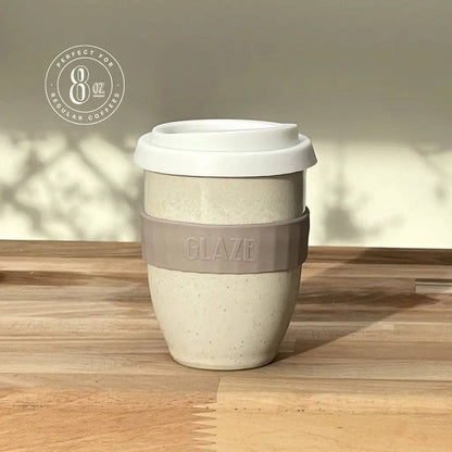 A reusable ceramic coffee cup with a white lid and a taupe-gray silicone sleeve embossed with the brand name 'GLAZE'. The cup has a speckled, warm off-white ceramic body.