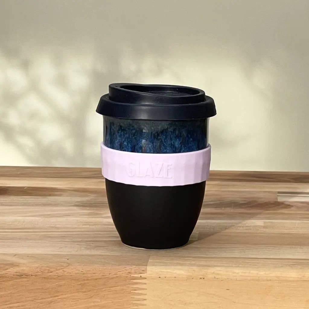 A reusable travel tumbler with a black lid and black lower body, a dark blue marbled upper section, and a light pink silicone sleeve wrapped around the middle. The sleeve has the brand name 'SLAZE' embossed on it.