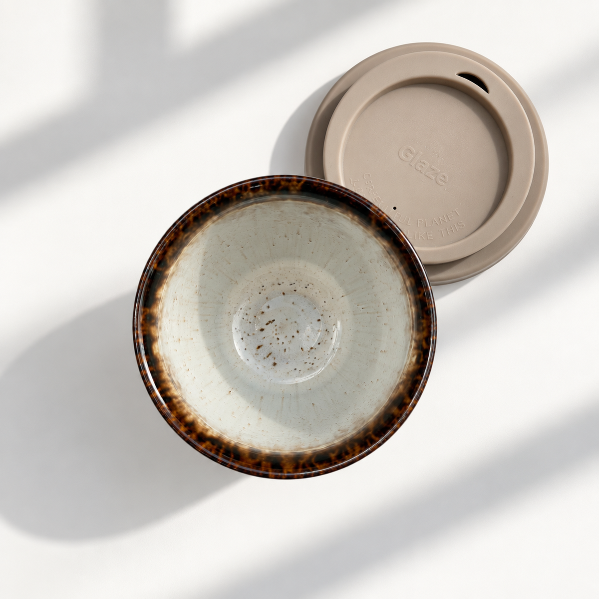 A ceramic travel tumbler with a speckled cream interior and a dark brown mottled rim, shown with a matching beige plastic lid on a white surface with soft window shadows.