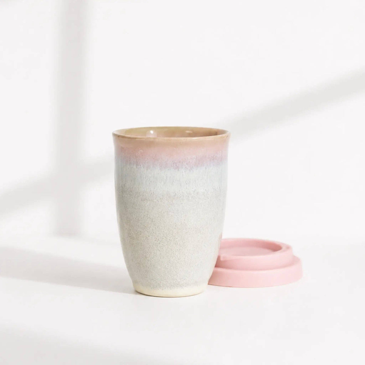 A ceramic travel mug with a pink and beige gradient glaze, shown next to its matching pink lid on a clean white surface.