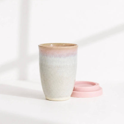 A ceramic travel mug with a pink and beige gradient glaze, shown next to its matching pink lid on a clean white surface.