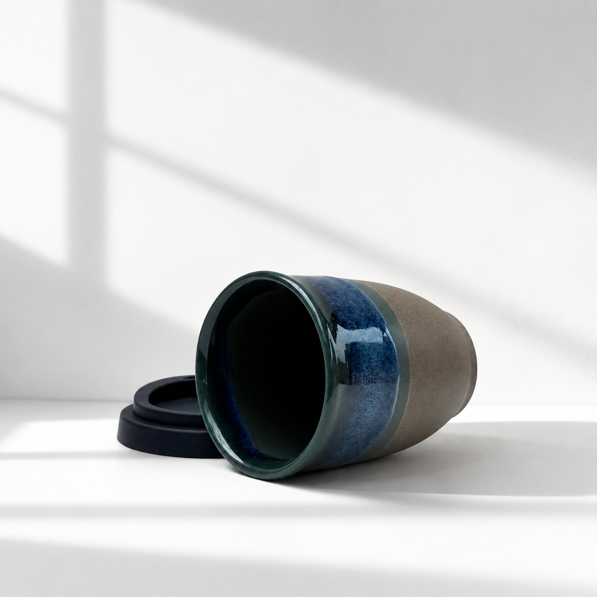 A ceramic travel tumbler with a dark blue glazed exterior and a matte taupe-brown textured body, shown lying on its side with a black silicone sleeve next to it on a white surface with soft window shadow patterns.