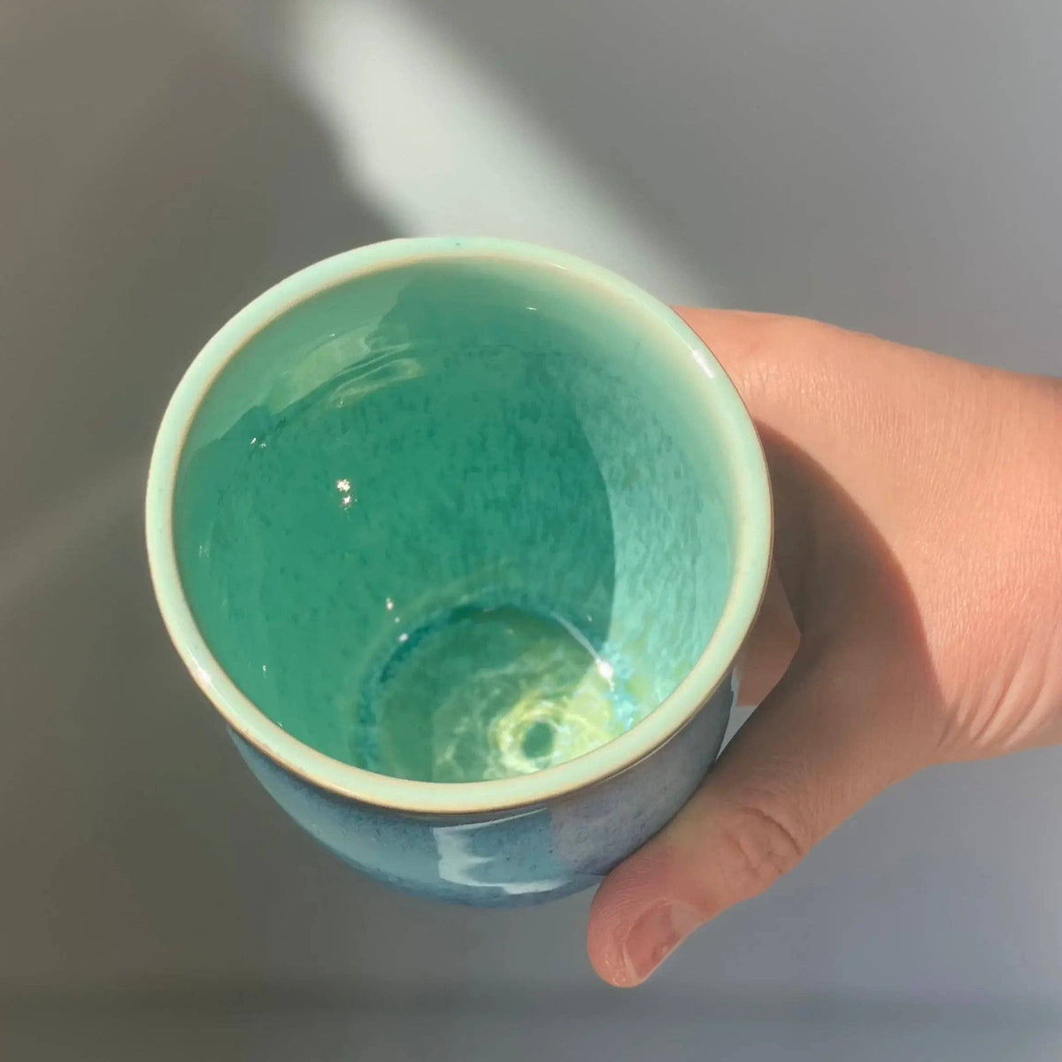 A hand holding a ceramic cup with a glossy turquoise and light blue glaze, photographed against a soft gray background with natural light.