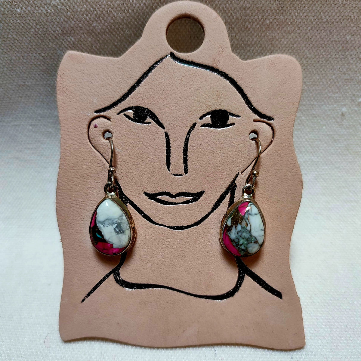 A pair of dangling earrings with teardrop-shaped marbled gemstone pendants in silver-toned settings, displayed on a tan leather card with a simple face illustration.