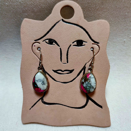 A pair of dangling earrings with teardrop-shaped marbled gemstone pendants in silver-toned settings, displayed on a tan leather card with a simple face illustration.