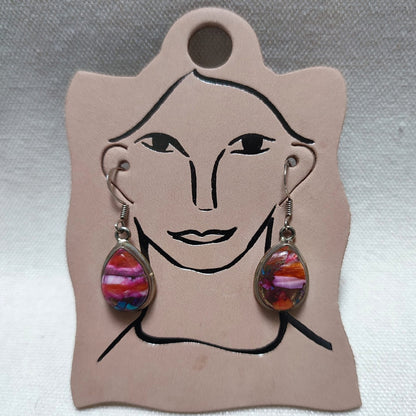 A pair of dangling teardrop earrings with colorful marbled pink, purple, orange, and blue glass inserts in silver-toned settings, displayed on a beige card with a simple face outline.