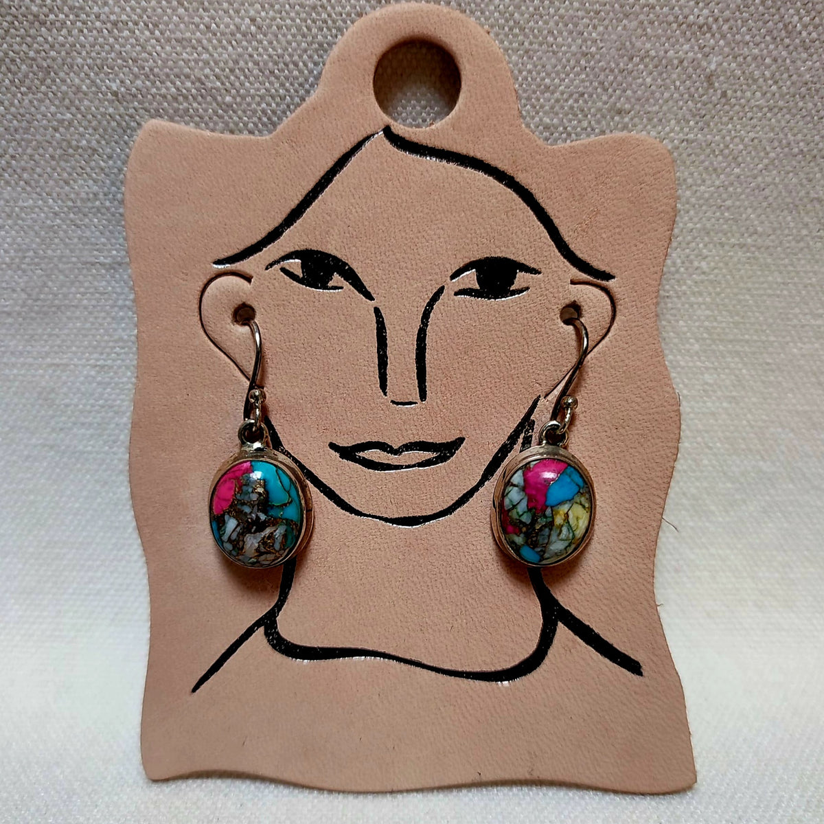 A pair of dangling earrings displayed on a tan leather card with a simple face outline. Each earring has a silver-toned hook and an oval cabochon featuring a colorful abstract design with pink, blue, green, and yellow tones.