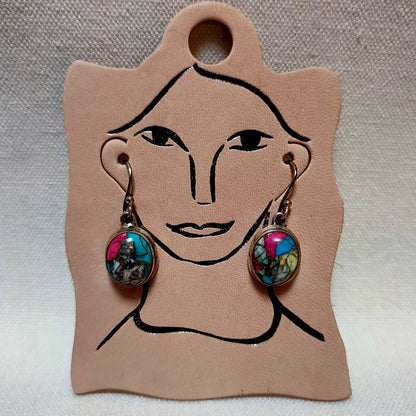 A pair of dangling earrings displayed on a tan leather card with a simple face outline. Each earring has a silver-toned hook and an oval cabochon featuring a colorful abstract design with pink, blue, green, and yellow tones.