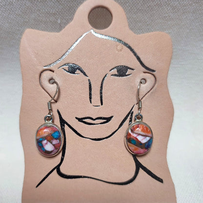 A pair of dangling earrings displayed on a tan card shaped like a stylized face. The earrings have silver-toned wire hooks and oval cabochons filled with a multicolored marbled pattern in orange, blue, pink, and white.