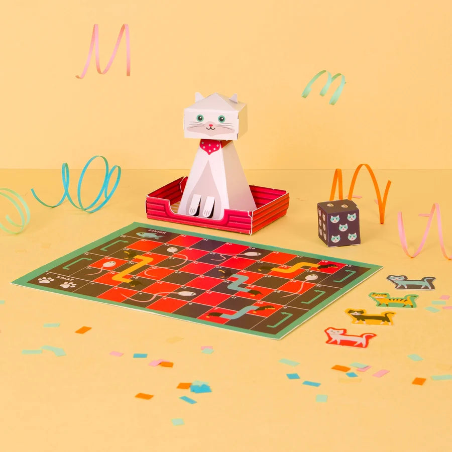 A colorful cat-themed board game set displayed on a warm yellow background. The set includes a square game board with illustrated tracks, a white cat figure sitting in a red basket, a dark dice cube with cat faces, and several small cat-shaped tokens scattered around.