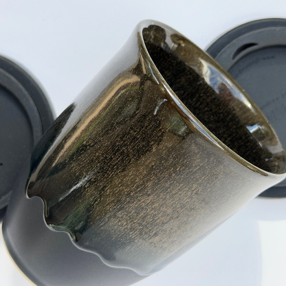 A close-up of a glossy ceramic cup with a dark brown and black gradient glaze, shown on a white background with part of a matching dark lid visible.